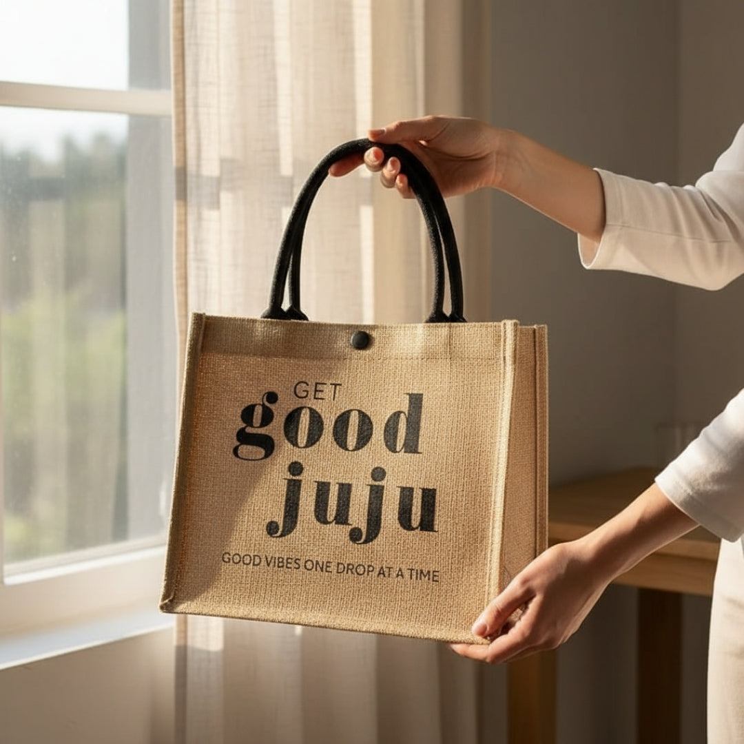 Two hands holding up a small natural jute tote bag with black handles, printed with the text “Get Good Juju” and “Good Vibes One Drop at a Time,” resting on a light-colored surface against a neutral indoor background.