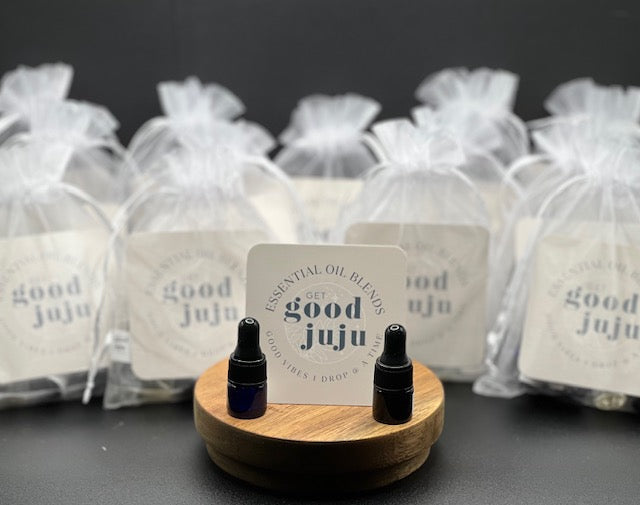 Two small essential oil sample bottles displayed on a round wooden stand in front of a Get Good Juju essential oil blends card, with multiple white organza sample bags labeled “good juju” that contain 6 small samples in them arranged in the background on a dark surface.