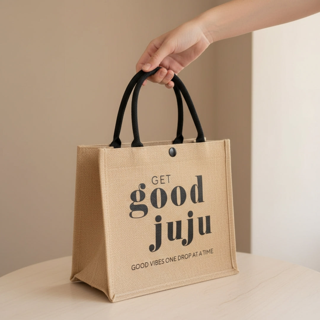 A hand holding a small natural jute tote bag with black handles, printed with the text “Get Good Juju” and “Good Vibes One Drop at a Time,” resting on a light-colored surface against a neutral indoor background.
