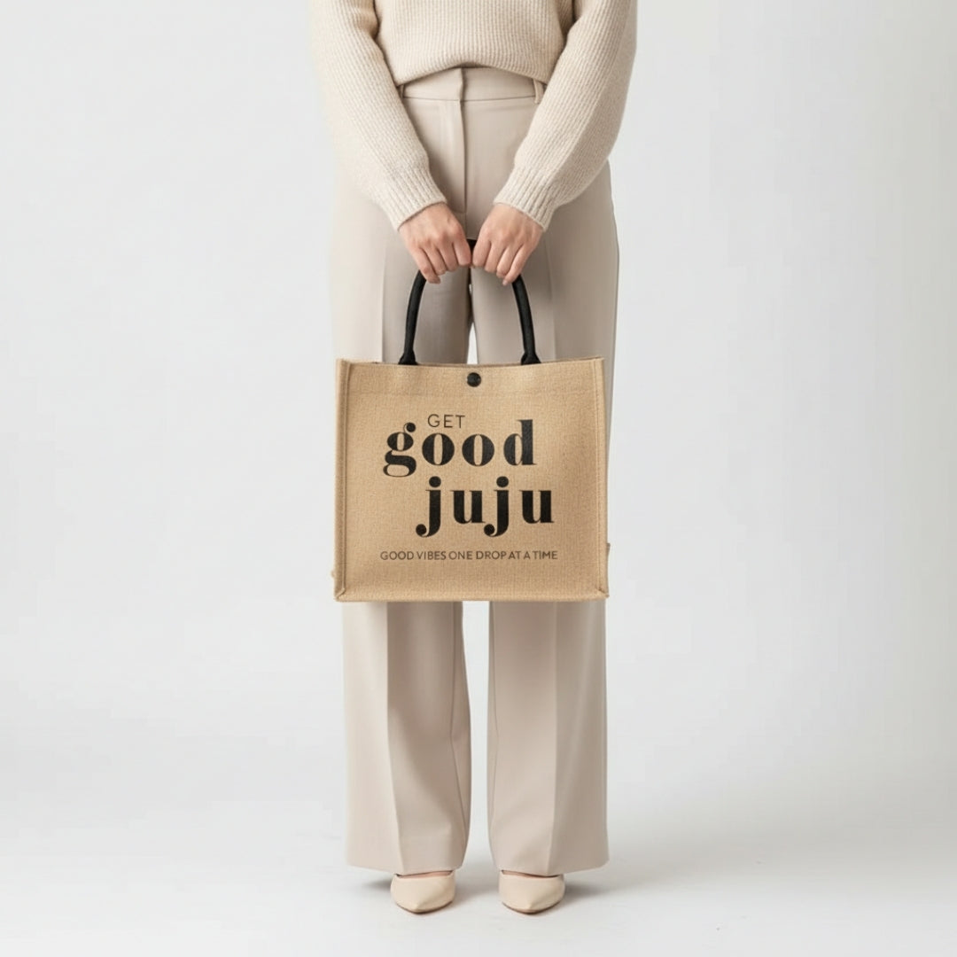 A person standing against a plain light background holding a natural jute tote bag with black handles, printed with the text “Get Good Juju” and “Good Vibes One Drop at a Time,” while wearing neutral-toned clothing and shoes.